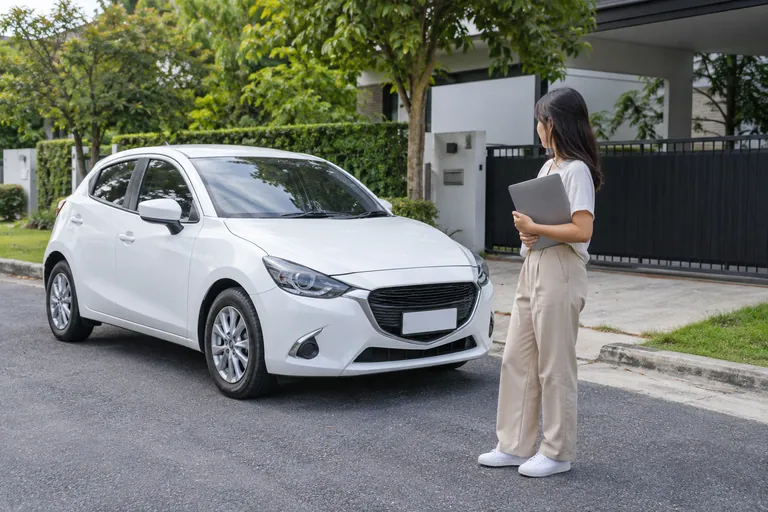Wanita memegang laptop tertutup bersebelahan hatchback putih di kawasan kediaman berturap.